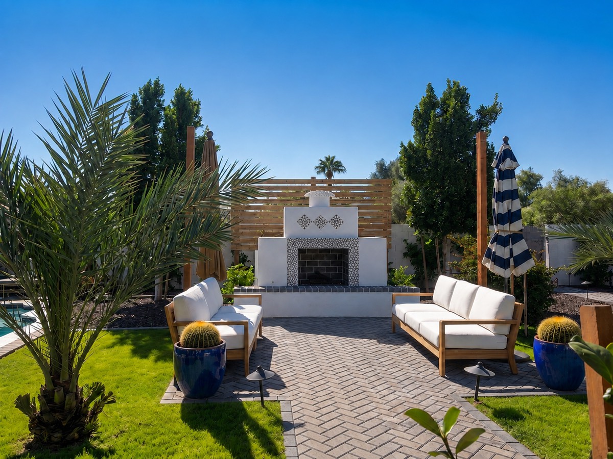 A desert backyard terrace in daylight: an outdoor fireplace, teak seating, potted barrel cactus and a date palm.