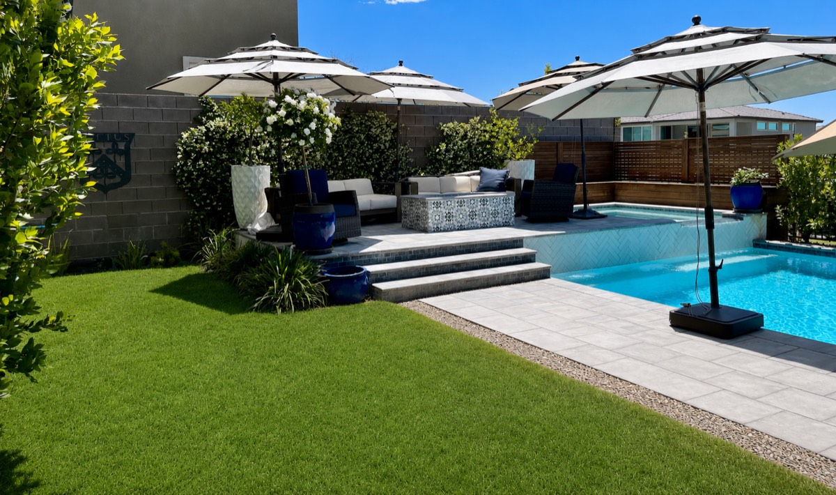 A Henderson, Nevada backyard photographed in daylight: pool, paved terrace, umbrellas and lawn.